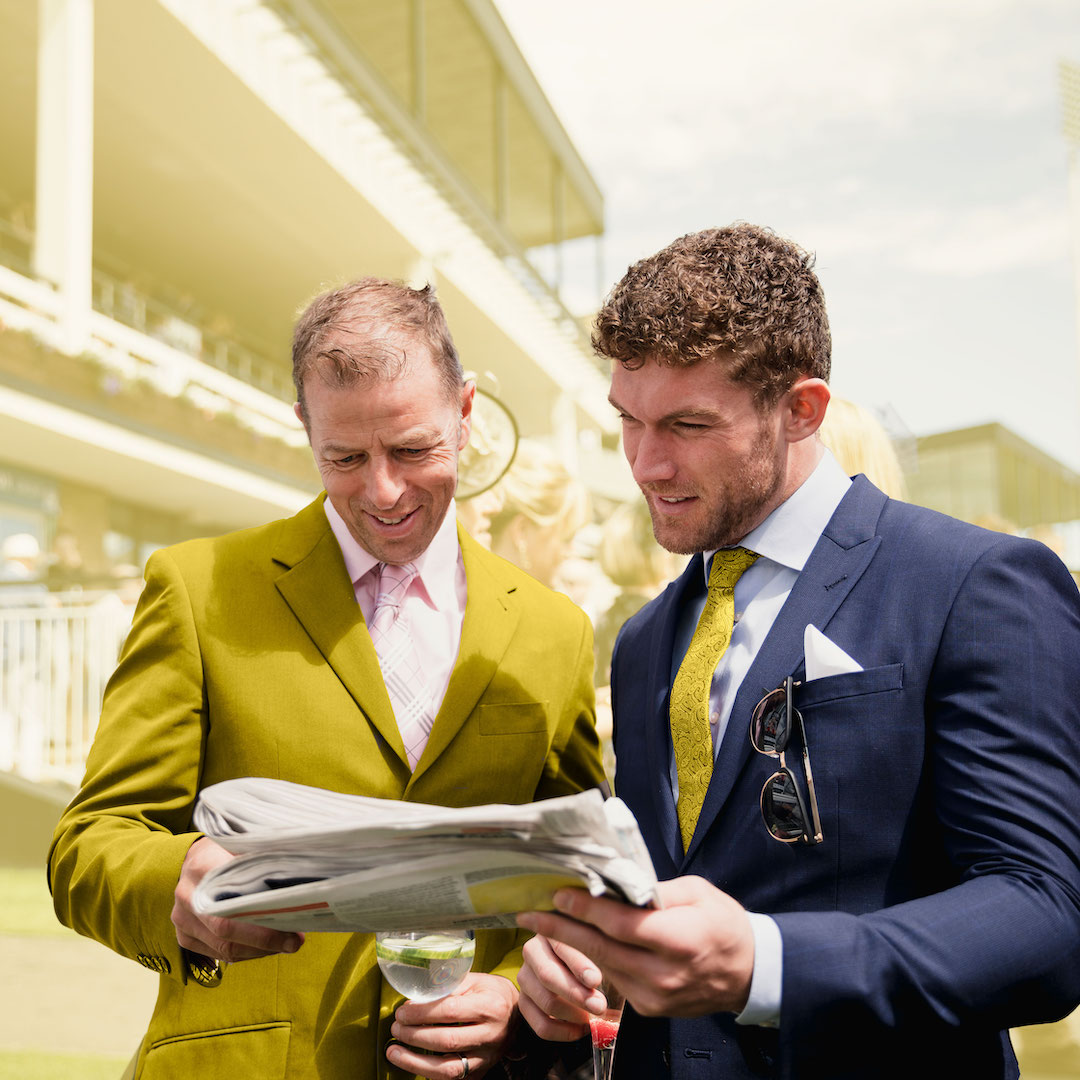 Two men at a horse race track inspecting the form